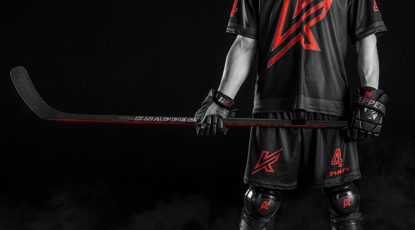 Close-up of a black and red ball hockey stick on a black background, with a player wearing ball hockey equipment.